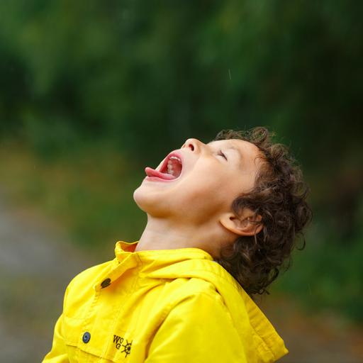 Ein Kind in gelber Regenjacke legt den Kopf in den Nacken und streckt mit weit geöffnetem Mund die Zunge heraus.