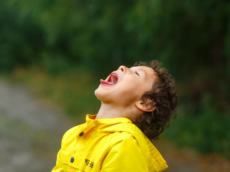 Ein Kind in gelber Regenjacke legt den Kopf in den Nacken und streckt mit weit geöffnetem Mund die Zunge heraus.