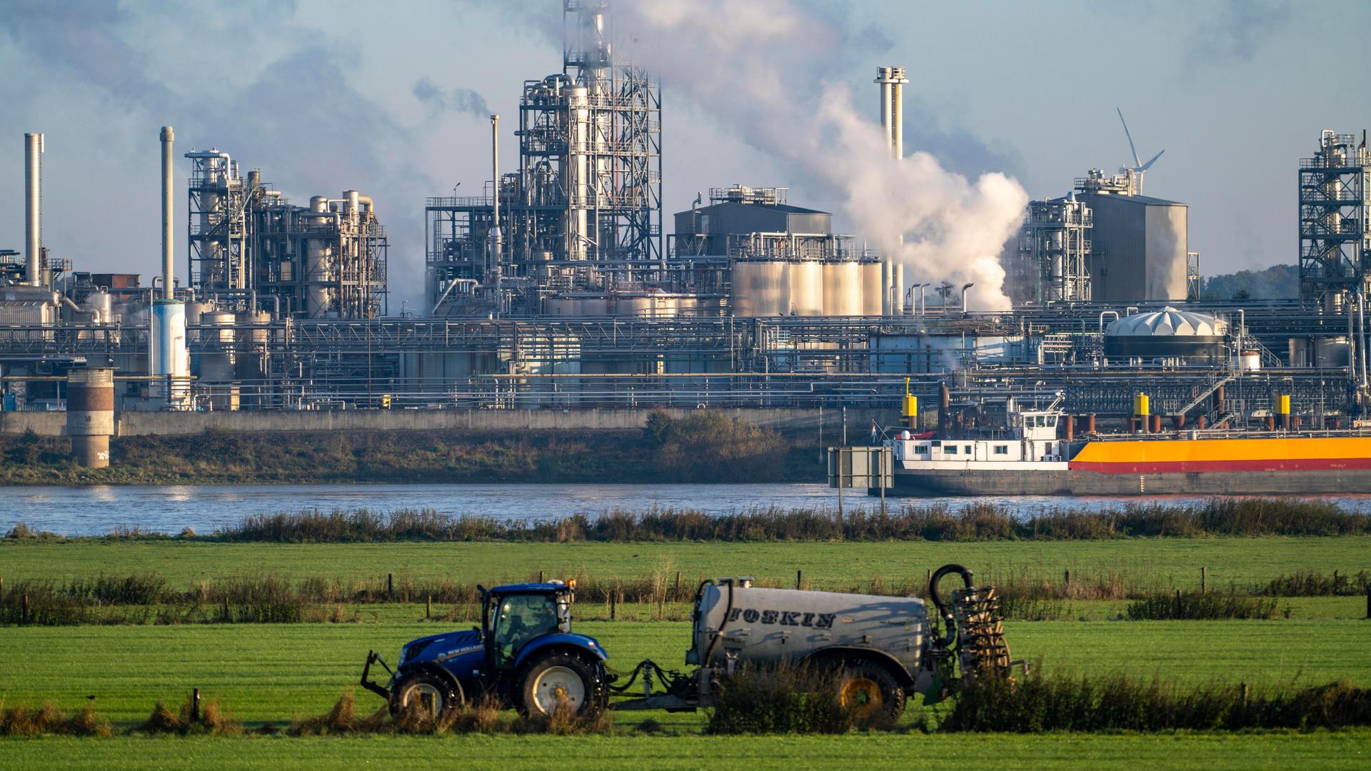 Traktor bringt auf einem Feld neben dem Rhein auf Höhe von Emmerich in Nordrhein-Westfalen Gülle aus. Auf dem Fluss fährt ein Frachtschiff, dahinter ist ein Industriebetrieb zu sehen. 