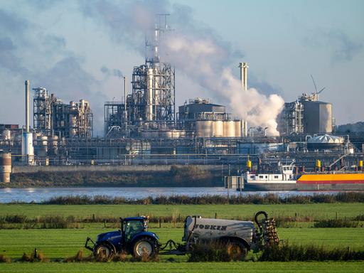 Traktor bringt auf einem Feld neben dem Rhein auf Höhe von Emmerich in Nordrhein-Westfalen Gülle aus. Auf dem Fluss fährt ein Frachtschiff, dahinter ist ein Industriebetrieb zu sehen. 