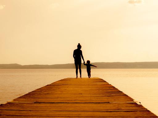 Eine Mutter mit kleinem Kind steht auf einem Holzsteg am Wasser. Beide sind von hinten nur als Silhouette zu erkennen.