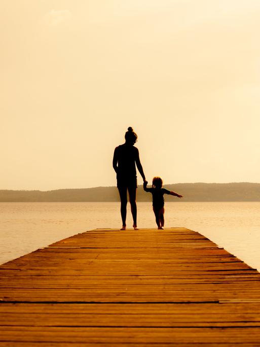 Eine Mutter mit kleinem Kind steht auf einem Holzsteg am Wasser. Beide sind von hinten nur als Silhouette zu erkennen.