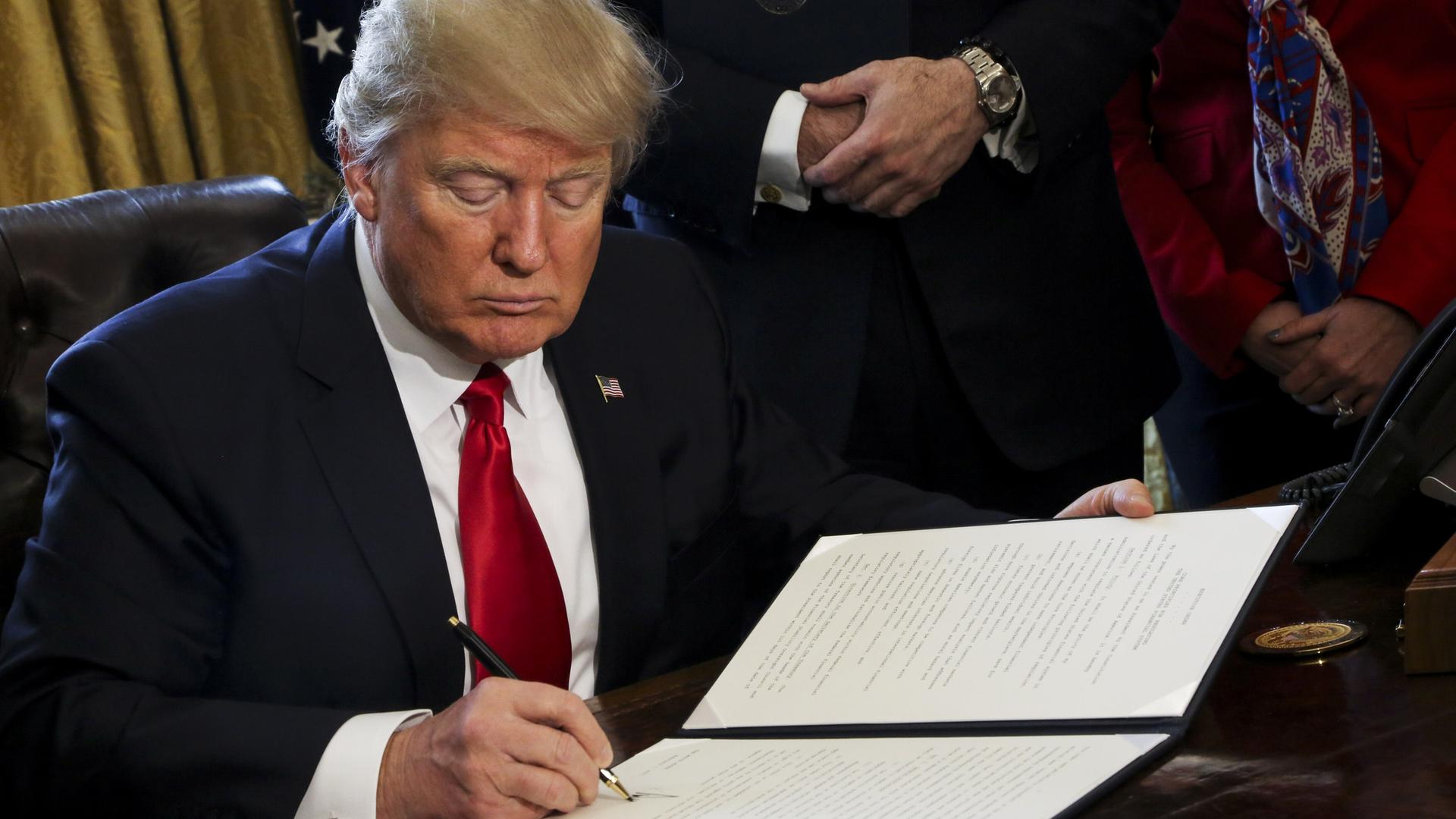 Der neue US-Präsident Trump sitzt am Schreibtisch in seinem Büro im Weißen Haus und unterzeichnet Dekrete, hinter ihm eine US-amerikanische Flagge