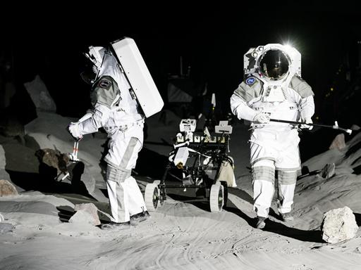 Zwei leere Astronautenanzüge sind auf einer künstlichen Mondlandschaft aufgestellt. Im Hintergrund: ein Mondfahrzeug. 