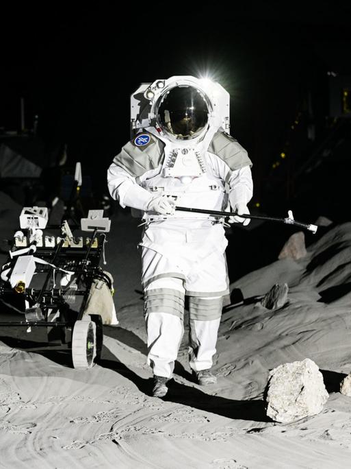 Zwei leere Astronautenanzüge sind auf einer künstlichen Mondlandschaft aufgestellt. Im Hintergrund: ein Mondfahrzeug. 