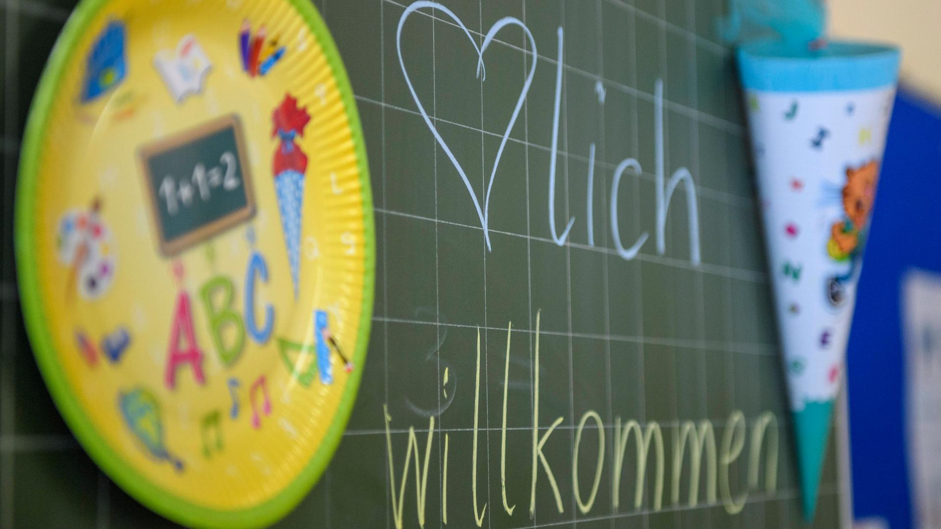 An der Tafel im Klassenzimmer einer Grundschule steht "Herzlich Willkommen"; daneben sind eine Schultüte und ein Pappteller mit Einschulungssymbolen wie "ABC" und Buntstiften.