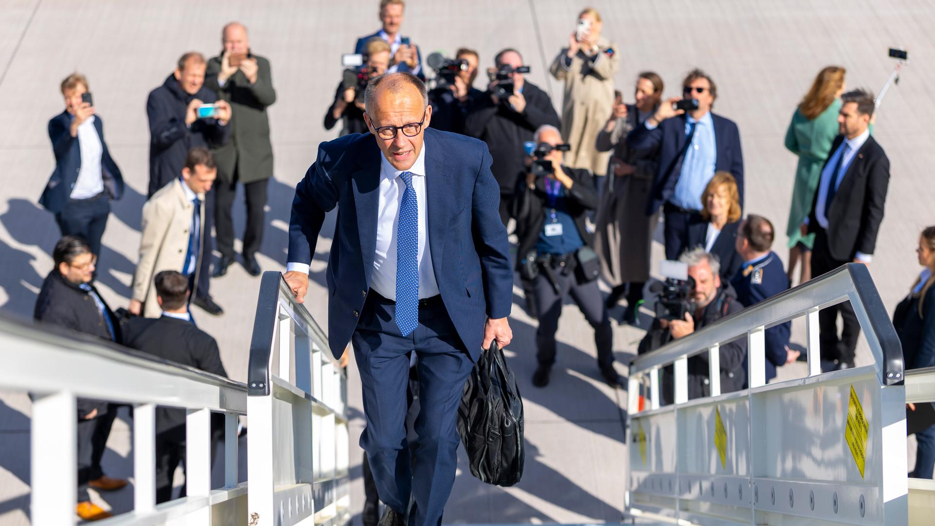 Friedrich Merz läuft die Gangway eines Fliegers hinauf. Unter ihm am Boden stehen Fotografen und fotografieren zu ihm hinauf.