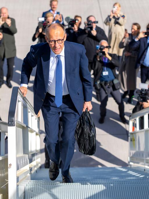 Friedrich Merz läuft die Gangway eines Fliegers hinauf. Unter ihm am Boden stehen Fotografen und fotografieren zu ihm hinauf.