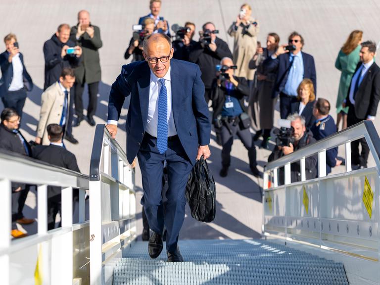 Friedrich Merz läuft die Gangway eines Fliegers hinauf. Unter ihm am Boden stehen Fotografen und fotografieren zu ihm hinauf.