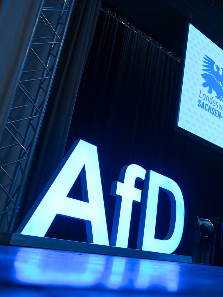 AfD-Schriftzug und das AfD-Logo auf der Bühne einem Versammlungssaal. 
