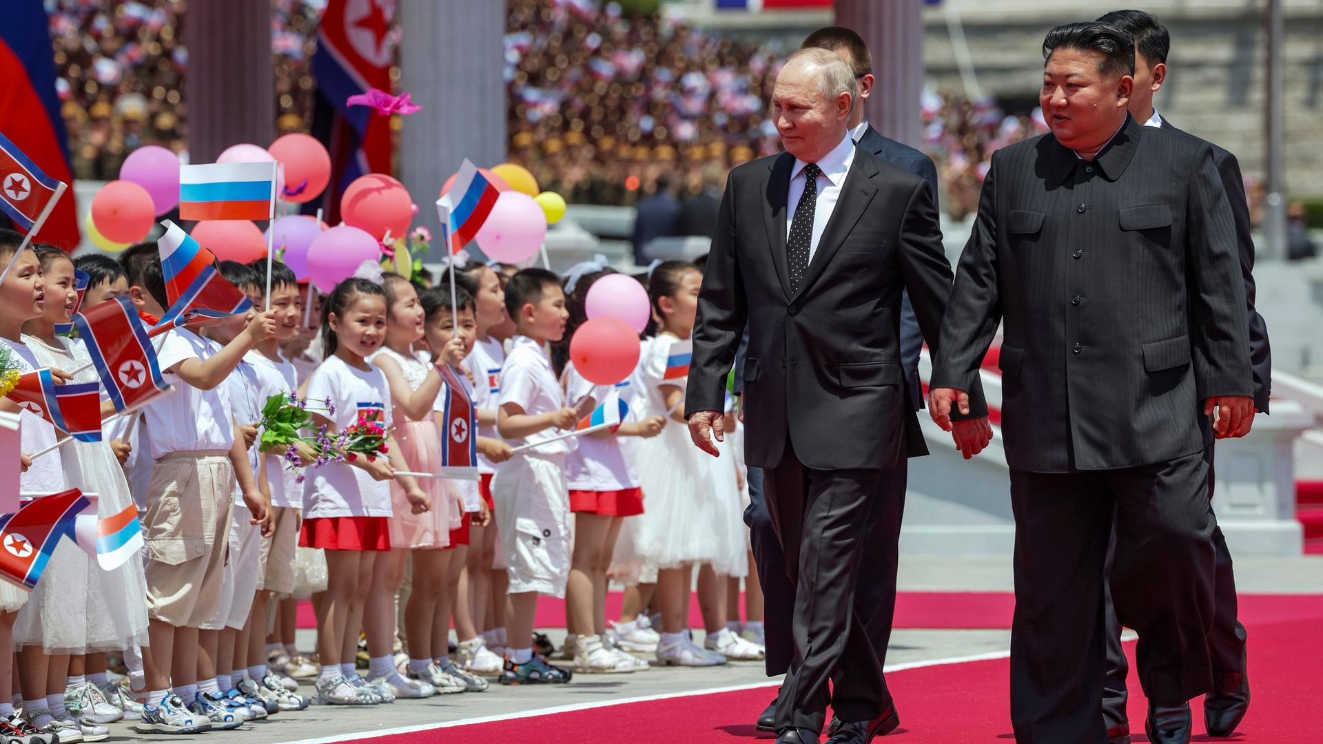 Der russische Präsident Wladimir Putin und Nordkoreas Machthaber Kim Jong Un gehen auf dem roten Teppich auf dem Kim Il Sung Platz in Pjöngjang. Hinter ihnen sind Kinder mit Fahnen und Luftballons, die ihnen zujubeln. Der russische Präsident Wladimir Putin und Nordkoreas Machthaber Kim Jong Un gehen auf dem roten Teppich auf dem Kim Il Sung Platz in Pjöngjang. Hinter ihnen sind Kinder mit Fahnen und Luftballons, die ihnen zujubeln.