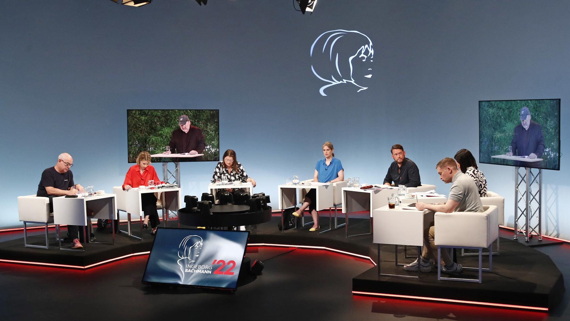 Ein Fernsehstudio mit mehreren sitzenden Juroren an Tischen; zwei Bildschirme zeigen einen Mann, der vorliest. 