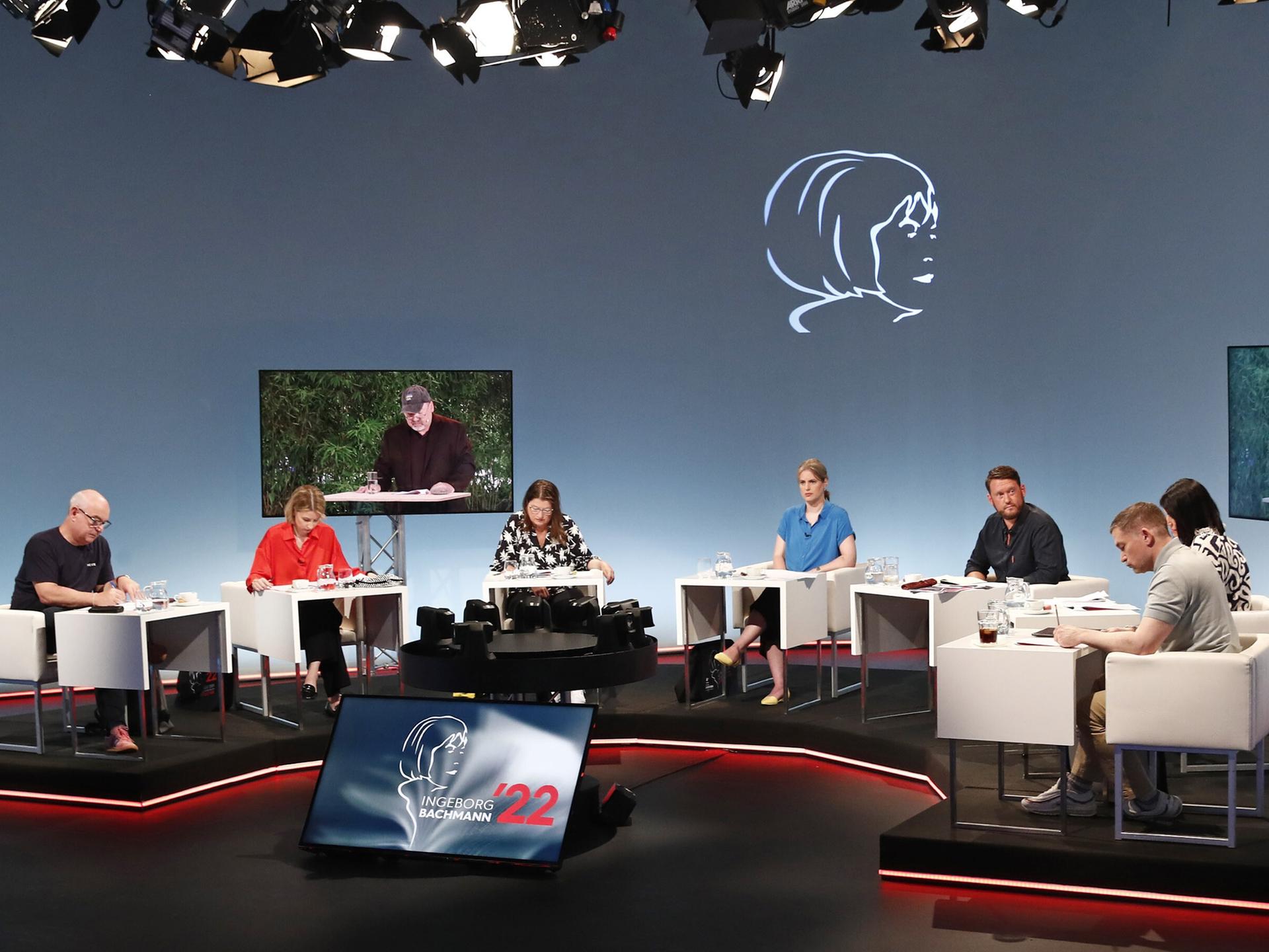 Ein Fernsehstudio mit mehreren sitzenden Juroren an Tischen; zwei Bildschirme zeigen einen Mann, der vorliest. 