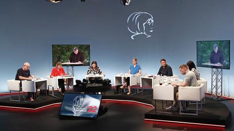 Ein Fernsehstudio mit mehreren sitzenden Juroren an Tischen; zwei Bildschirme zeigen einen Mann, der vorliest. 