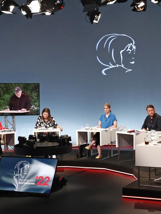 Ein Fernsehstudio mit mehreren sitzenden Juroren an Tischen; zwei Bildschirme zeigen einen Mann, der vorliest. 