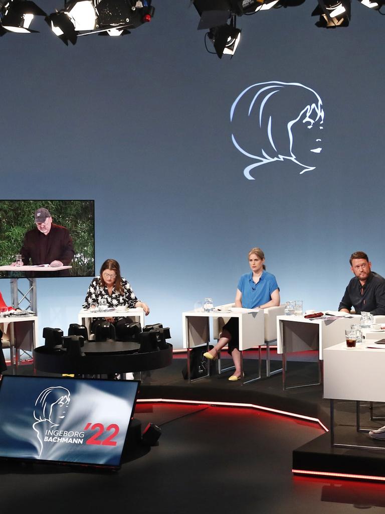 Ein Fernsehstudio mit mehreren sitzenden Juroren an Tischen; zwei Bildschirme zeigen einen Mann, der vorliest. 