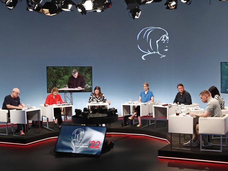 Ein Fernsehstudio mit mehreren sitzenden Juroren an Tischen; zwei Bildschirme zeigen einen Mann, der vorliest. 