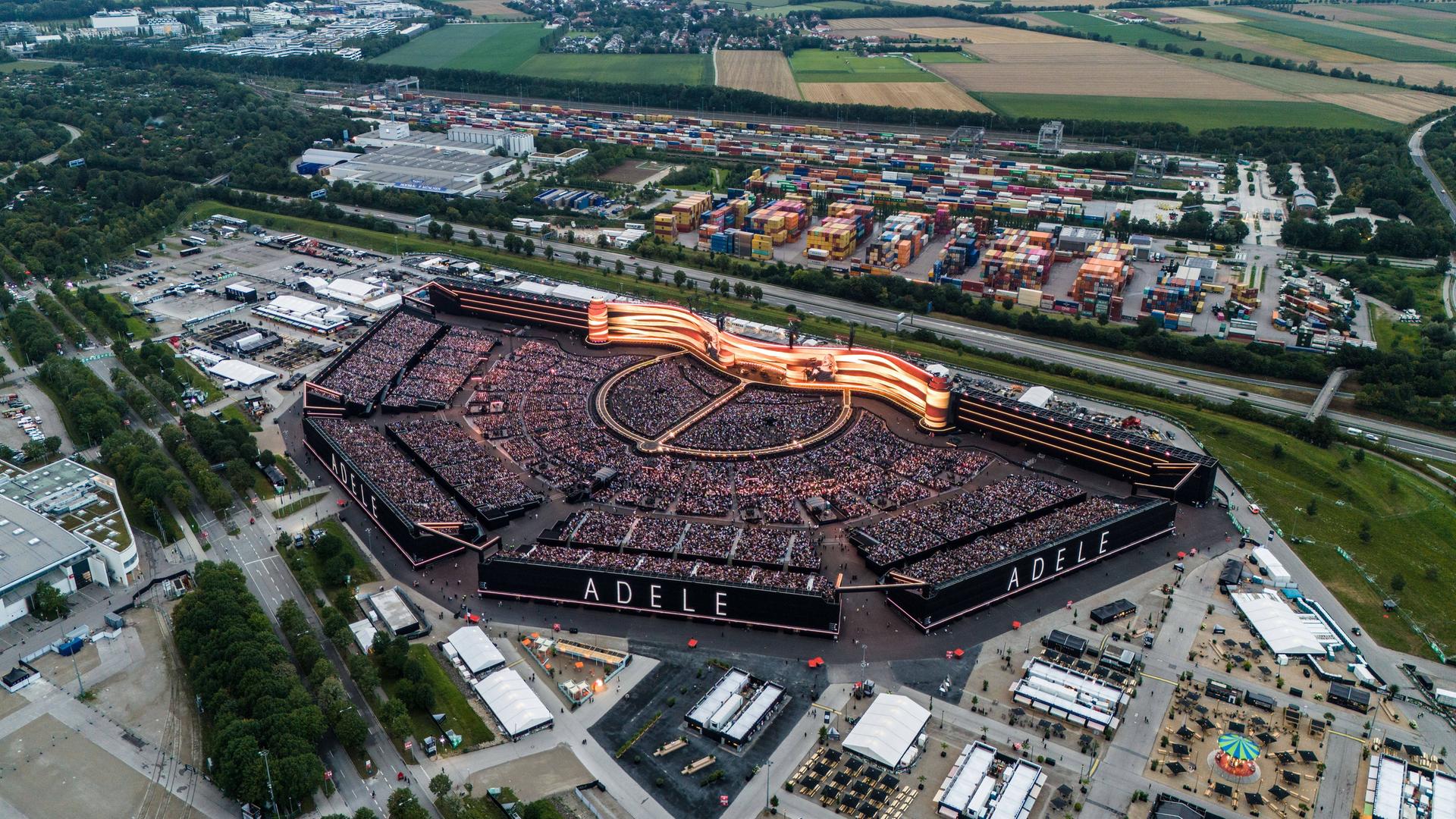 Ein riesiges Freiluft-Stadion mit dem Schriftzug Adele. Im Stadion ist eine gigantische, gebogene Leinwand zu sehen. 