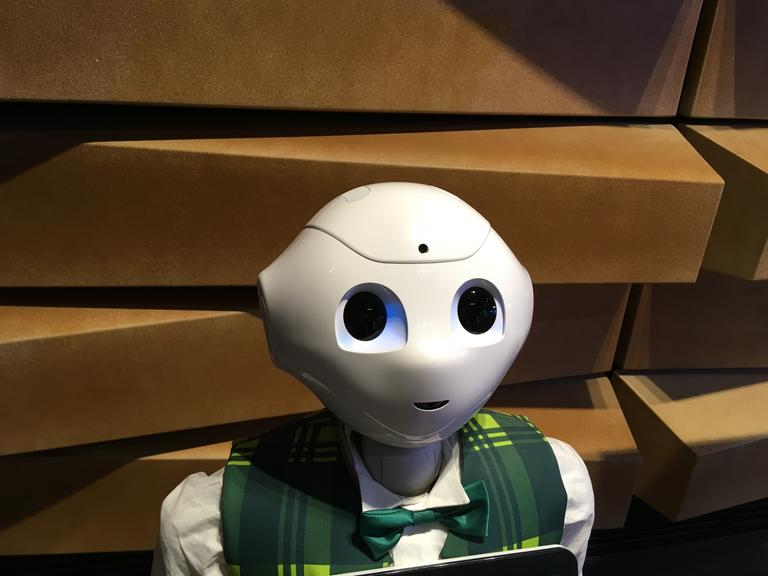 Kopf und Schultern des humanoiden Roboters "Pepper": glatte weiße Oberfläche, sehr große Augen, eine angedeutete Nase und ein kleiner lächelnder Mund. Er trägt eine karierte Weste und eine Fliege.
