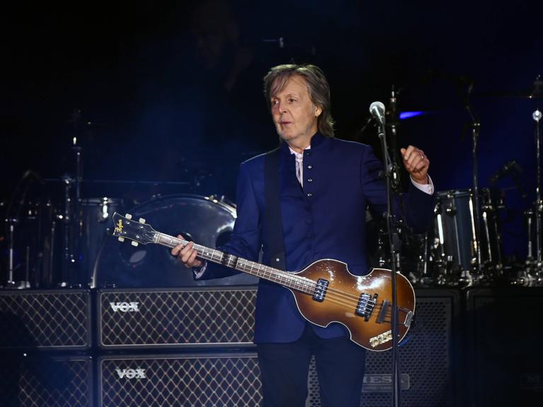 Ein Mann mit Gitarre steht auf einer Bühne. Er trägt eine dunkelblaue Jacke. Es ist der frühere Beatle Paul McCartney.