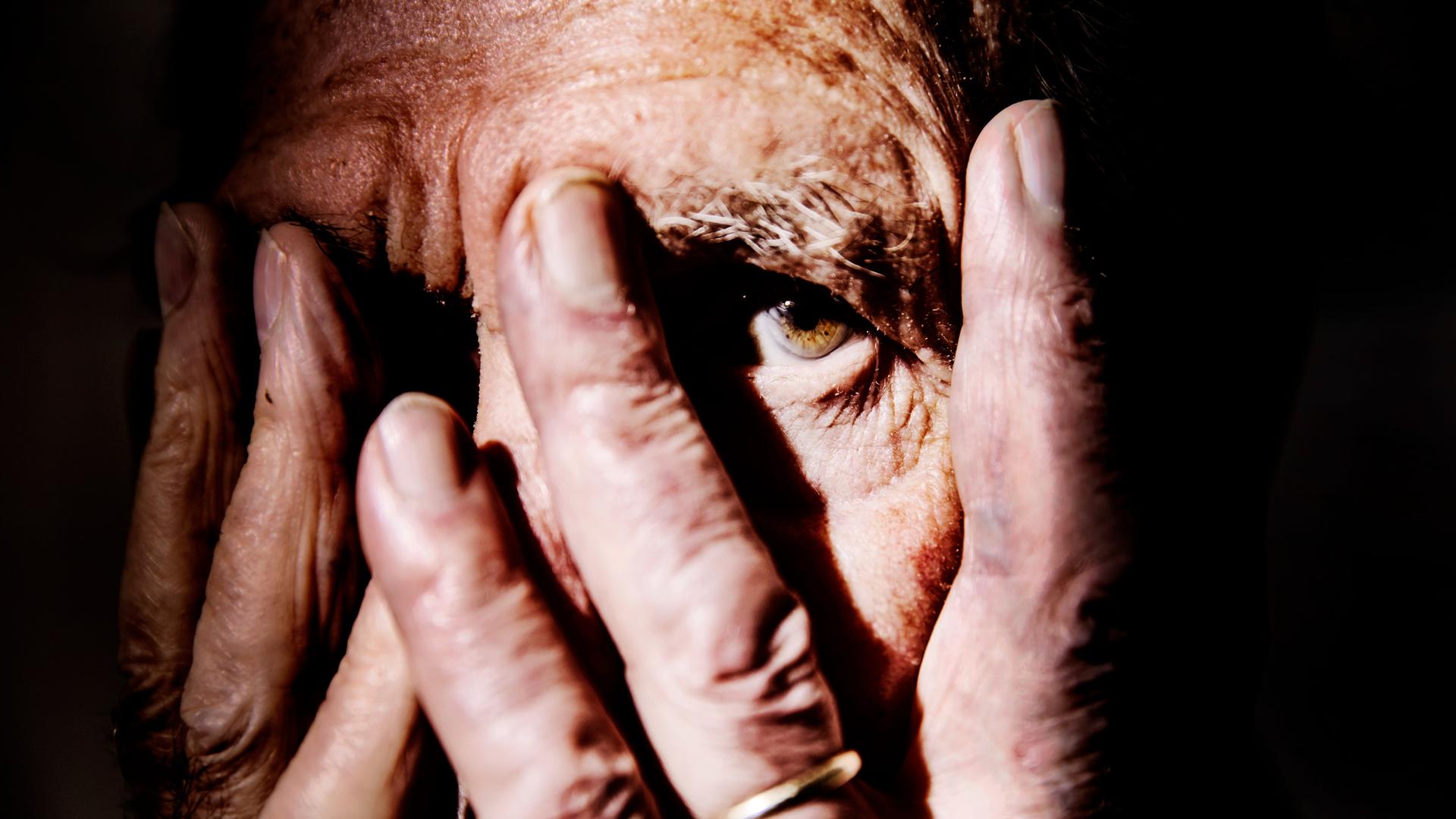 Nahaufnahme einer alten Person, die sich die Hände vor das Gesicht hält und mit einem Auge zwischen den Fingern hindurch schaut. Der Hintergrund ist schwarz.