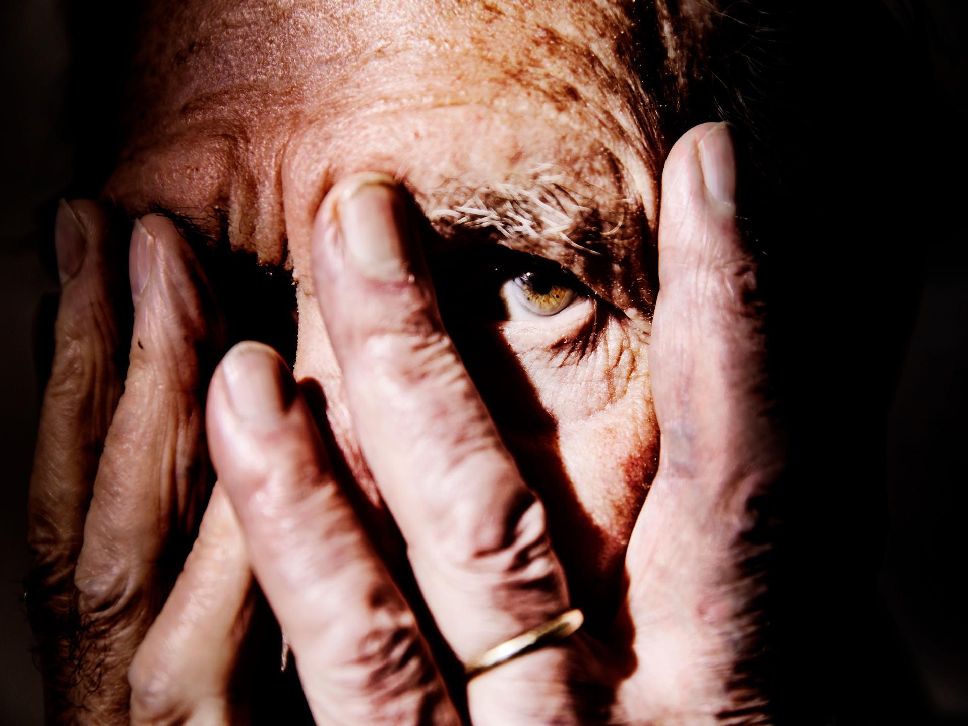 Nahaufnahme einer alten Person, die sich die Hände vor das Gesicht hält und mit einem Auge zwischen den Fingern hindurch schaut. Der Hintergrund ist schwarz. Nahaufnahme einer alten Person, die sich die Hände vor das Gesicht hält und mit einem Auge zwischen den Fingern hindurch schaut. Der Hintergrund ist schwarz.