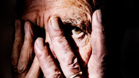 Nahaufnahme einer alten Person, die sich die Hände vor das Gesicht hält und mit einem Auge zwischen den Fingern hindurch schaut. Der Hintergrund ist schwarz. Nahaufnahme einer alten Person, die sich die Hände vor das Gesicht hält und mit einem Auge zwischen den Fingern hindurch schaut. Der Hintergrund ist schwarz.