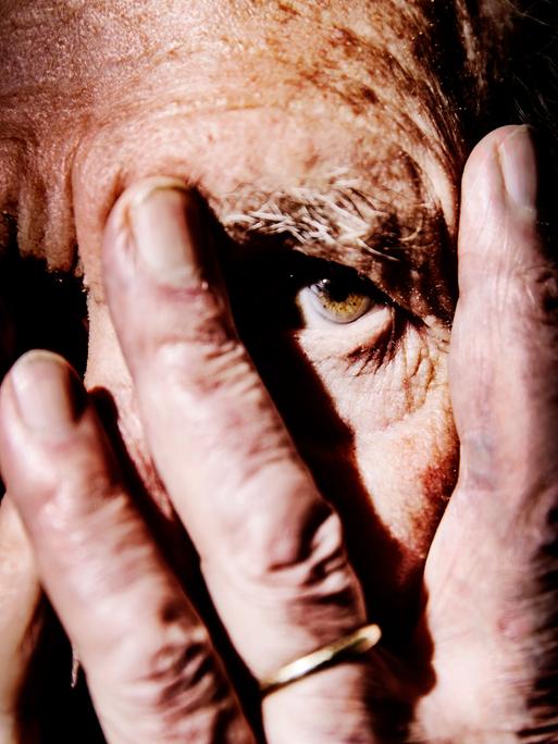 Nahaufnahme einer alten Person, die sich die Hände vor das Gesicht hält und mit einem Auge zwischen den Fingern hindurch schaut. Der Hintergrund ist schwarz.