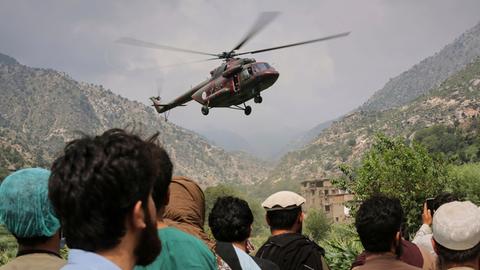 Ein Militärhubschrauber fliegt über Menschen in Afghanistan. Im Hintergrund sind Berge und Häuser zu sehen. 