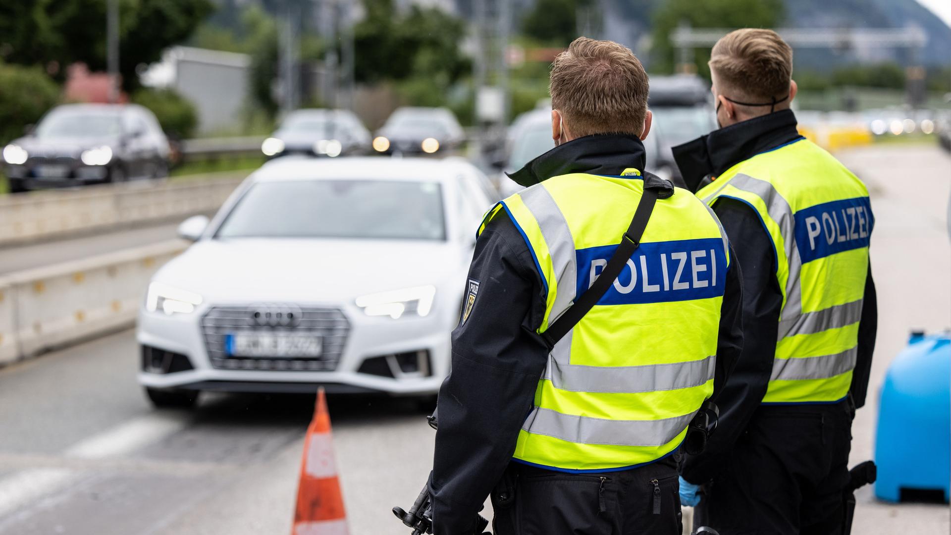 Zwei Bundespolizisten stehen an der deutsch-österreichischen Grenze und beobachten den vorbeifahrenden Verkehr. Man sieht sie von hinten. Beide tragen gelbe Signalwesten. Auf dem Rücken der beiden steht auf blauem Hintergrund in weiß "Polizei".