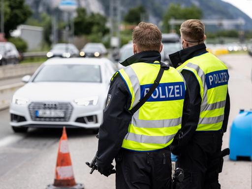 Zwei Bundespolizisten stehen an der deutsch-österreichischen Grenze und beobachten den vorbeifahrenden Verkehr. Man sieht sie von hinten. Beide tragen gelbe Signalwesten. Auf dem Rücken der beiden steht auf blauem Hintergrund in weiß "Polizei".