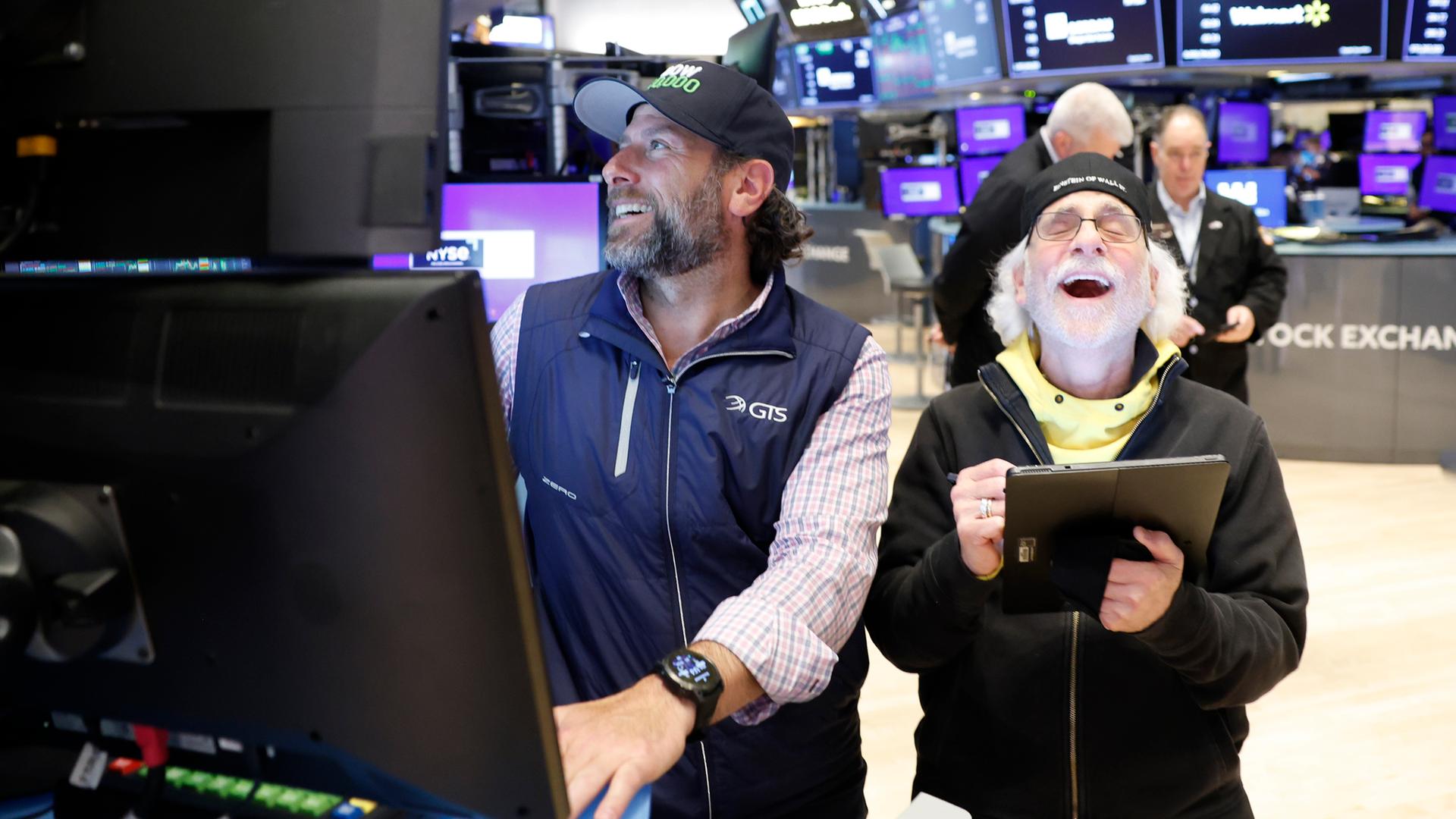 Zwei Bärtige Männer mit Baseballmützen stehen in einem Raum voller Bildschirme. Beide sind offenbar guter Dinge. Der rechte lacht mit weit geöffnetem Mund.