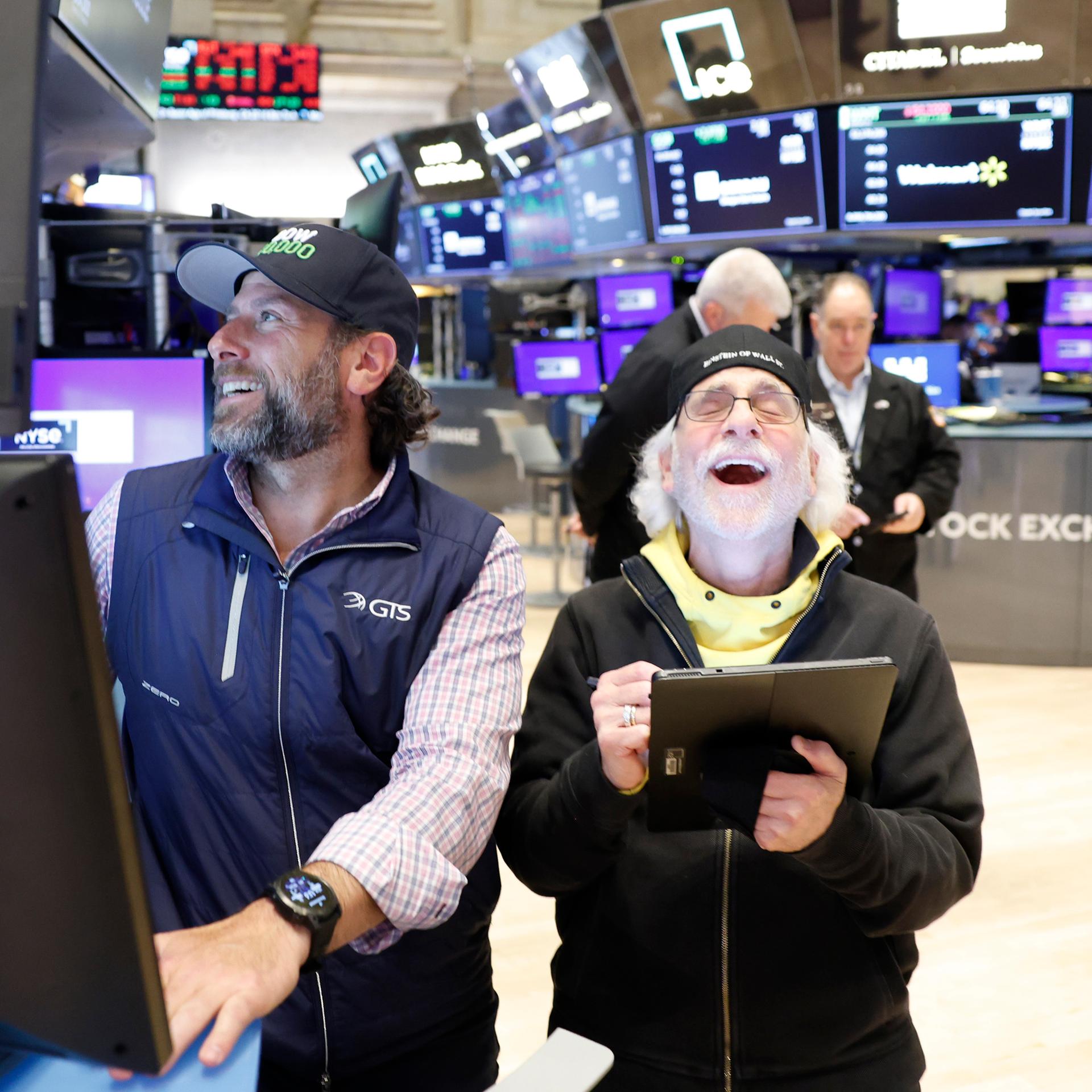 Zwei Bärtige Männer mit Baseballmützen stehen in einem Raum voller Bildschirme. Beide sind offenbar guter Dinge. Der rechte lacht mit weit geöffnetem Mund.