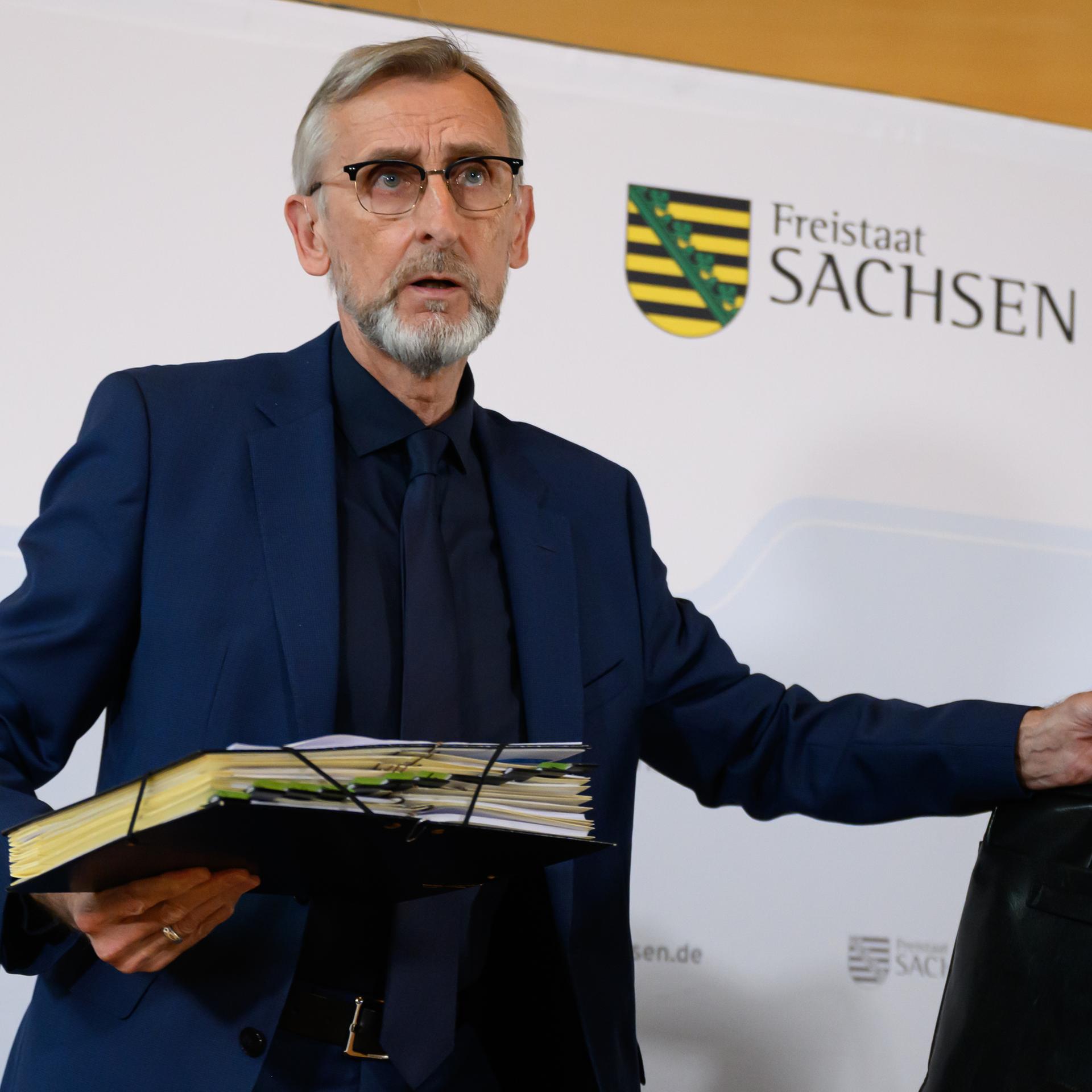 Armin Schuster (CDU), Innenminister in Sachsen, nimmt vor der Pressekonferenz zur Sitzung des sächsischen Kabinetts in der Offizierschule des Heeres im Scharnhorst-Saal auf dem Podium Platz und hält dabei eine Aktenmappe und eine Tasche in den Händen.