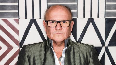 Ein Mann mit eine rechteckigen Brille steht vor einer Wand aus gemusterten Fliesen. Er blickt ernst.