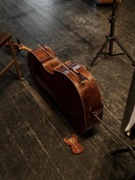 Ein Violoncello liegt neben einem Holzstuhl und einem Notenständer auf dem Boden, während der dazugehörige Bogen darauf abgelegt ist.