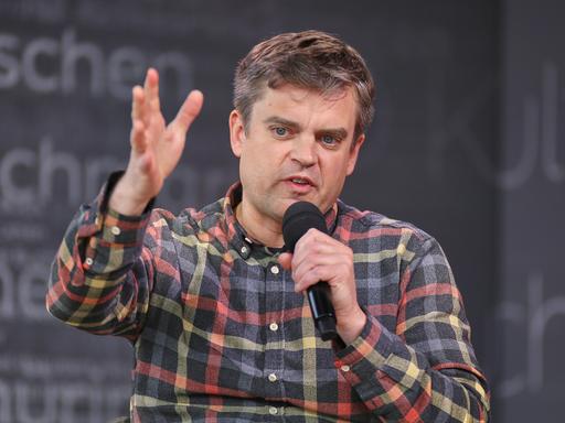 Matthias Nawrat sitzt in einem karierten Hemd in Leipzig, Sachsen, au der Bühne und spricht.