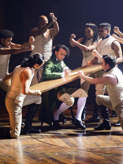 Szene aus dem Musical "Hamilton" im Operettenhaus Hamburg: Mehrere Tänzerinnen und Tänzer gruppieren sich um eine Holzplatte, die Benét Monteiro in der Hauptrolle als Schreibunterlage dient. 