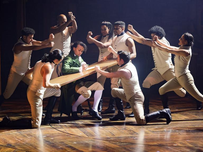 Szene aus dem Musical "Hamilton" im Operettenhaus Hamburg: Mehrere Tänzerinnen und Tänzer gruppieren sich um eine Holzplatte, die Benét Monteiro in der Hauptrolle als Schreibunterlage dient. 