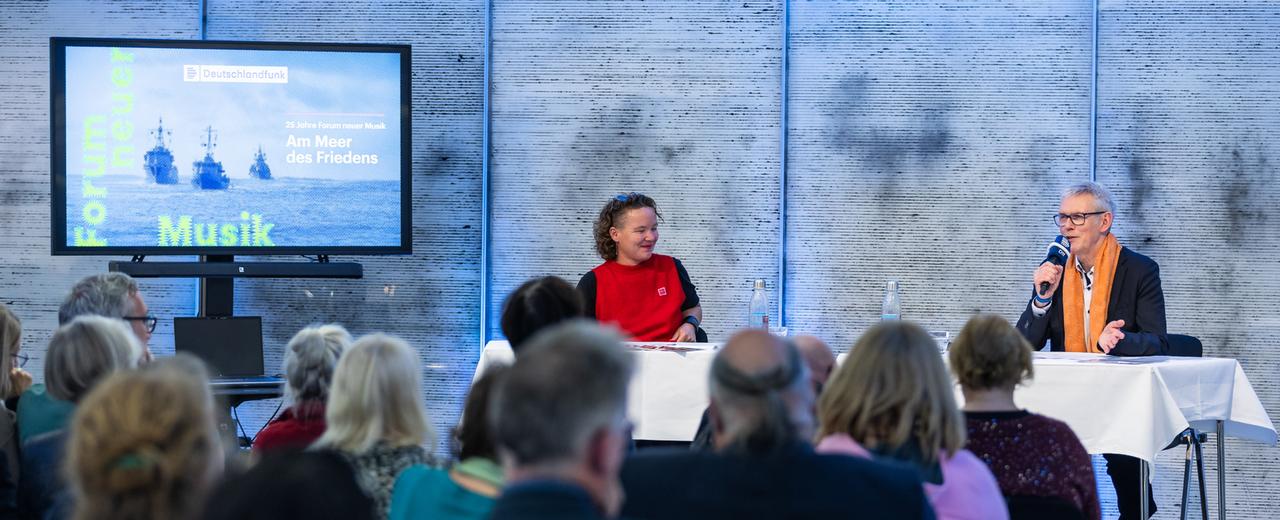 Bei einer Podiumsveranstaltung sitzen zwei Personen an einem Tisch vor einem Publikum.