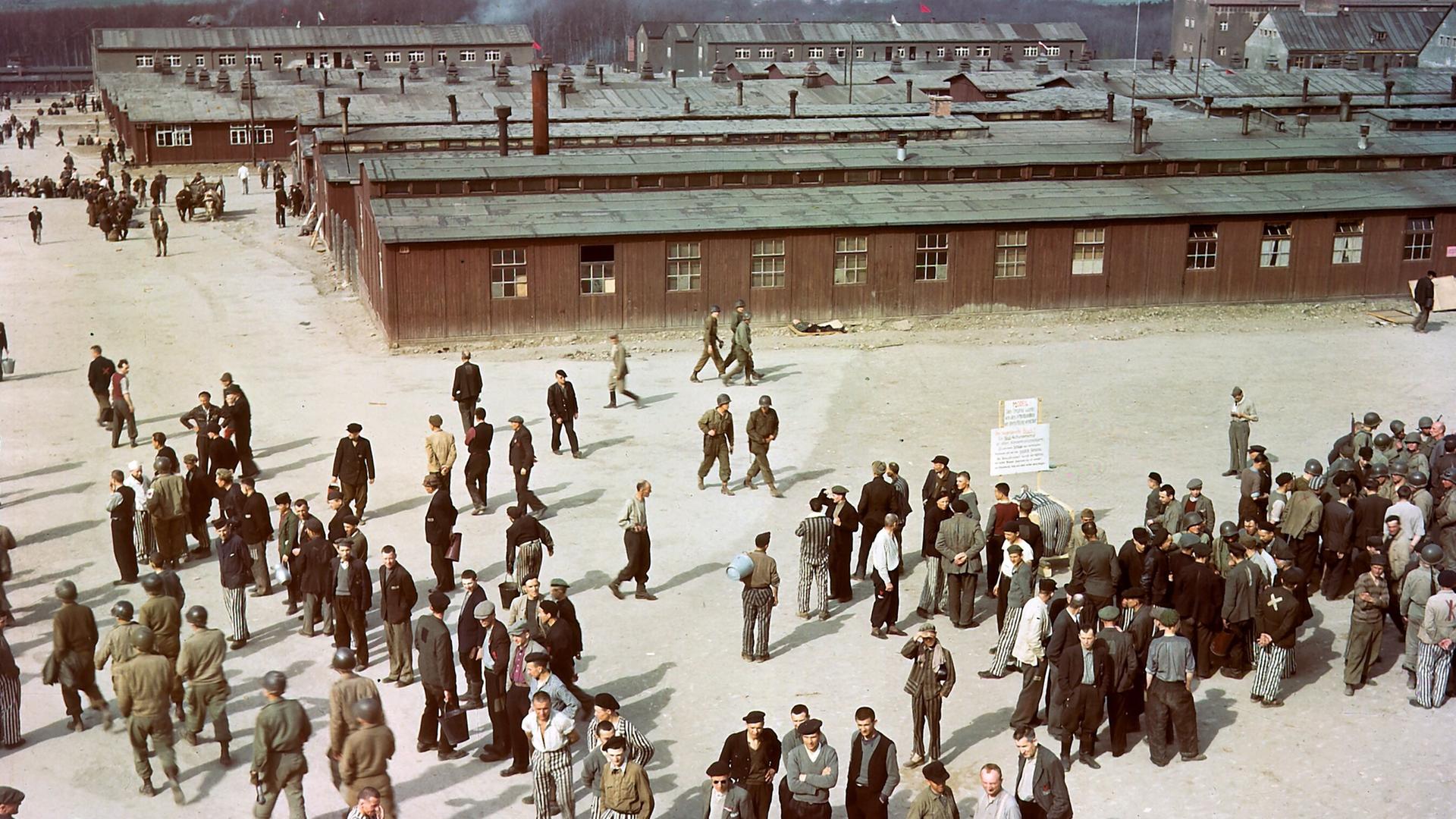 Luftaufnahme des Konzentrationslagers Buchenwald mit Baracken im Hintergrund. Auf einem Hof stehen ehemalige Häftlinge und US-Soldaten,