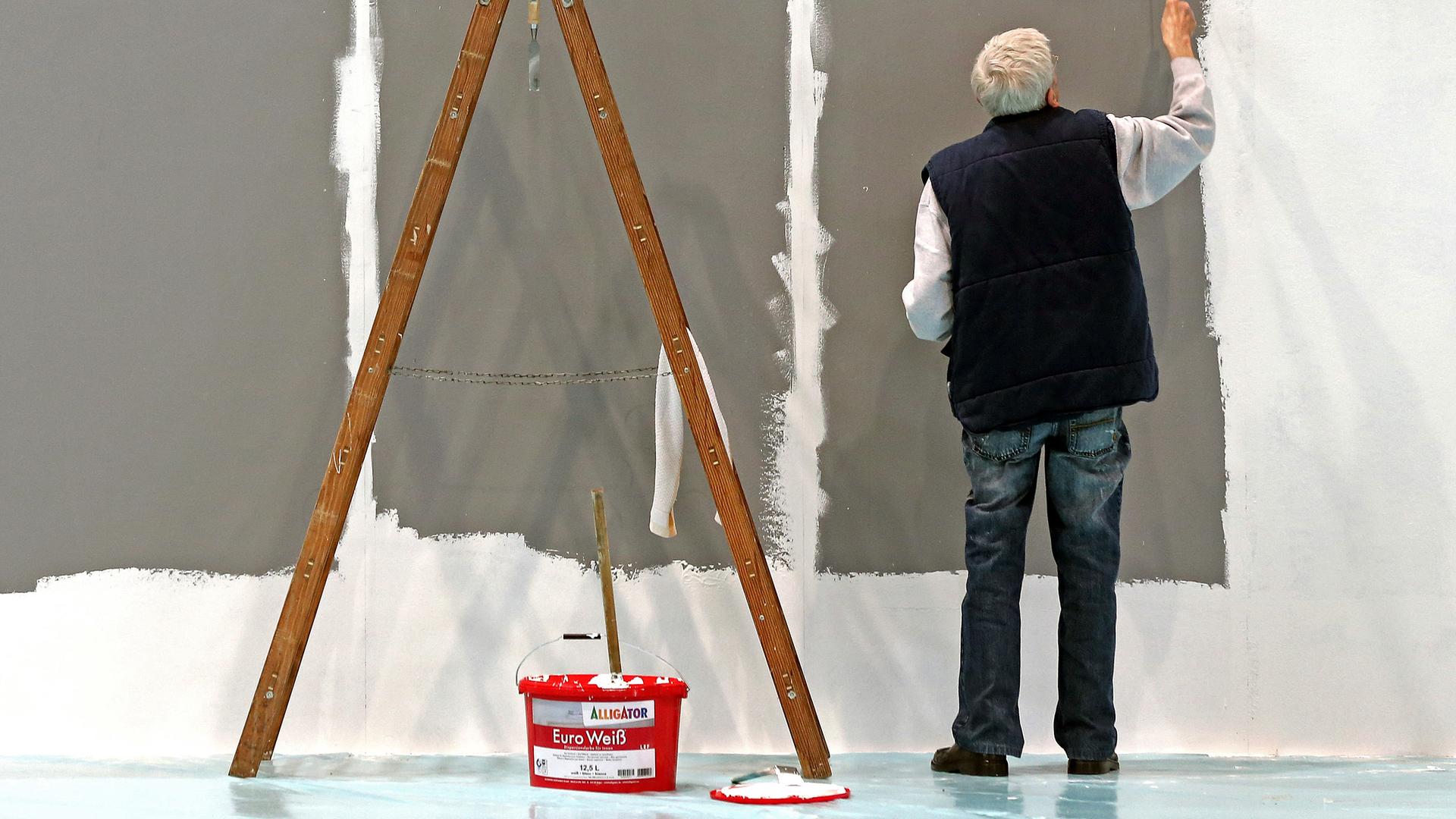 Ein älterer Mann mit grauen Haaren streicht eine Wand. 