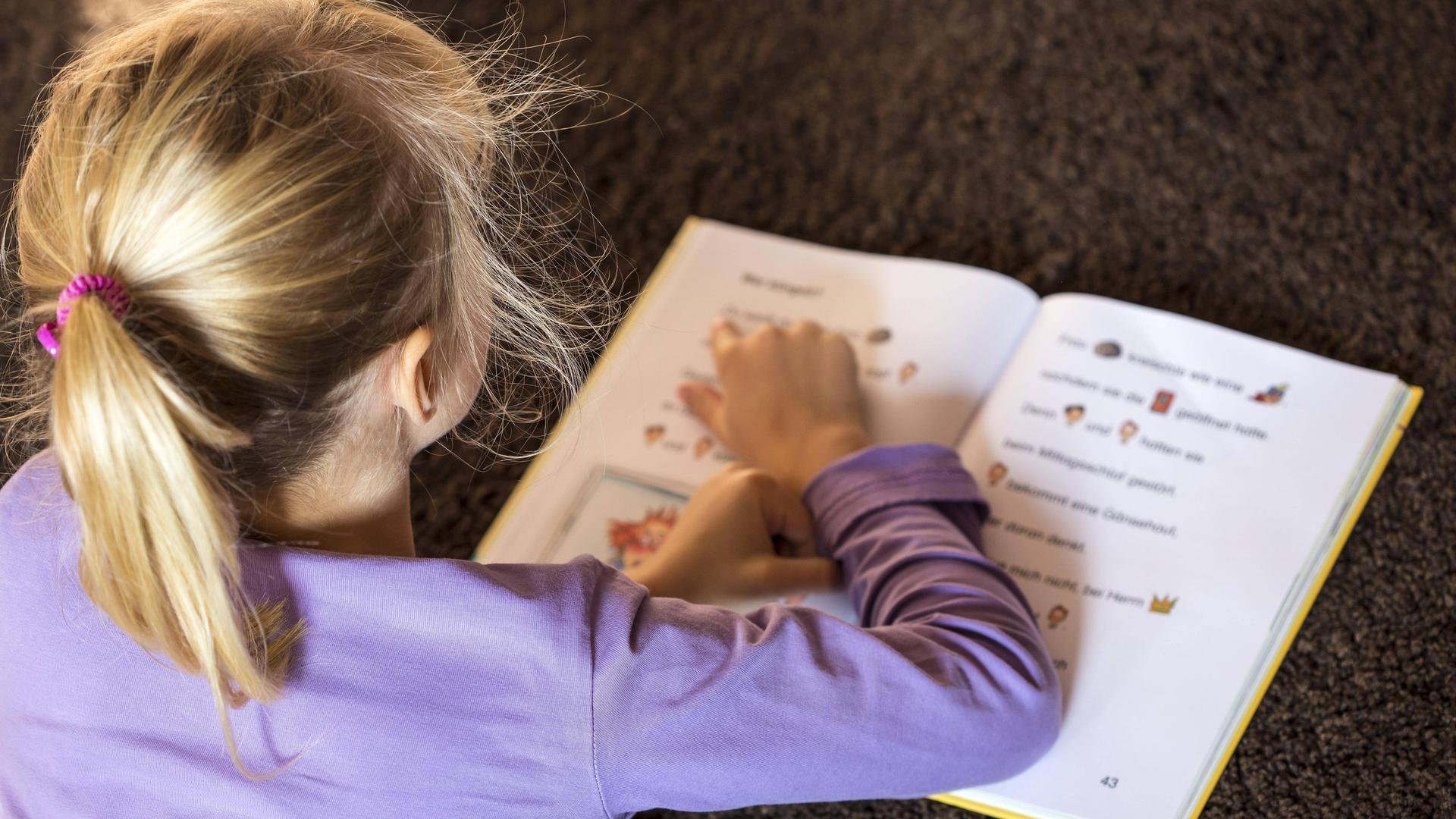 Ein Mädchen mit blonden Haaren liegt und liest ein Buch. Sie zeigt mit dem Finger auf die Buchstaben.
