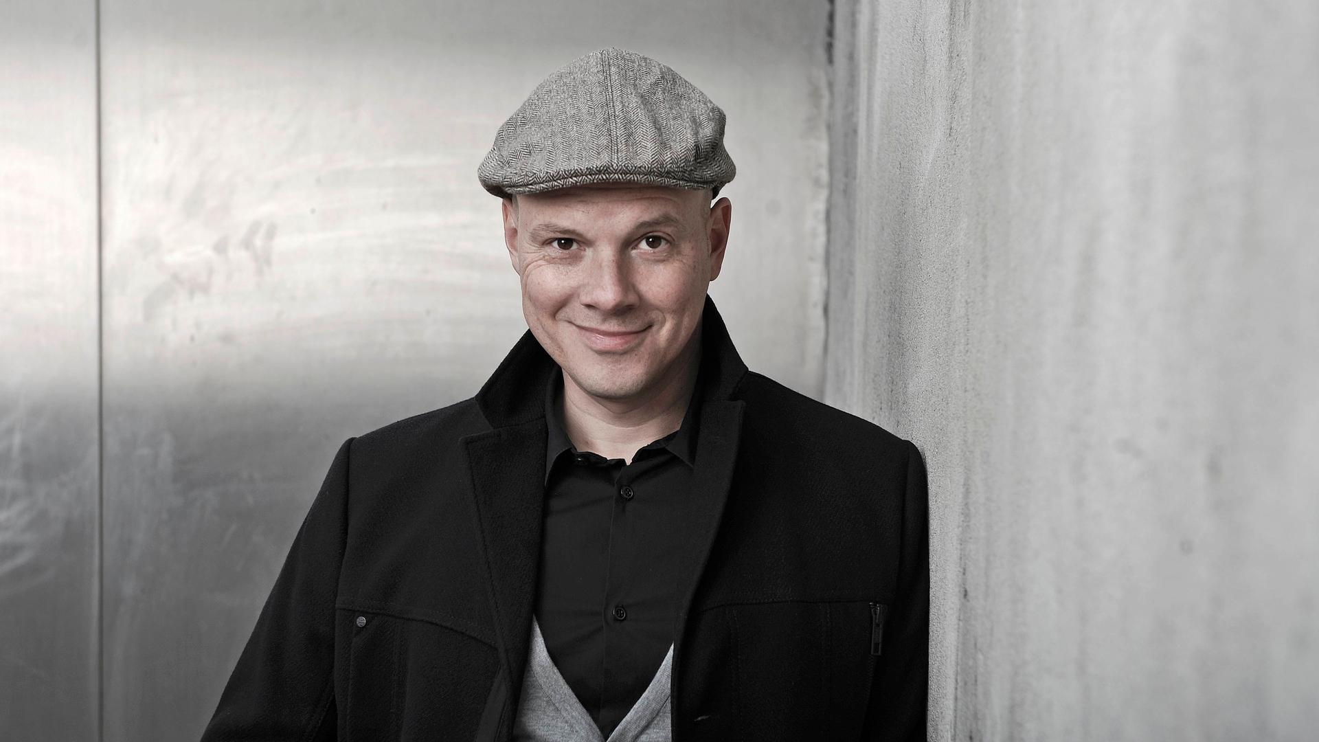 Oliver Hilmes. Er trägt eine schwarze Jacke, graue Schiebermütze und schaut lächelnd in die Kamera Oliver Hilmes. Er trägt eine schwarze Jacke, graue Schiebermütze und schaut lächelnd in die Kamera