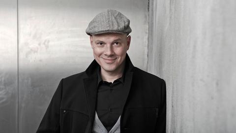 Oliver Hilmes. Er trägt eine schwarze Jacke, graue Schiebermütze und schaut lächelnd in die Kamera