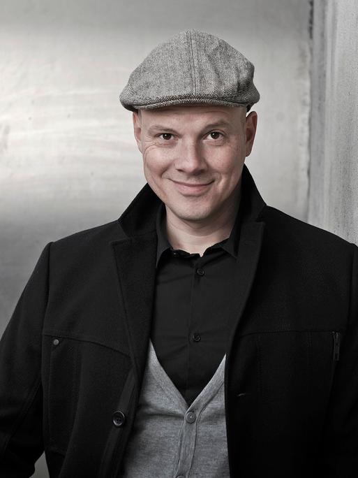 Oliver Hilmes. Er trägt eine schwarze Jacke, graue Schiebermütze und schaut lächelnd in die Kamera