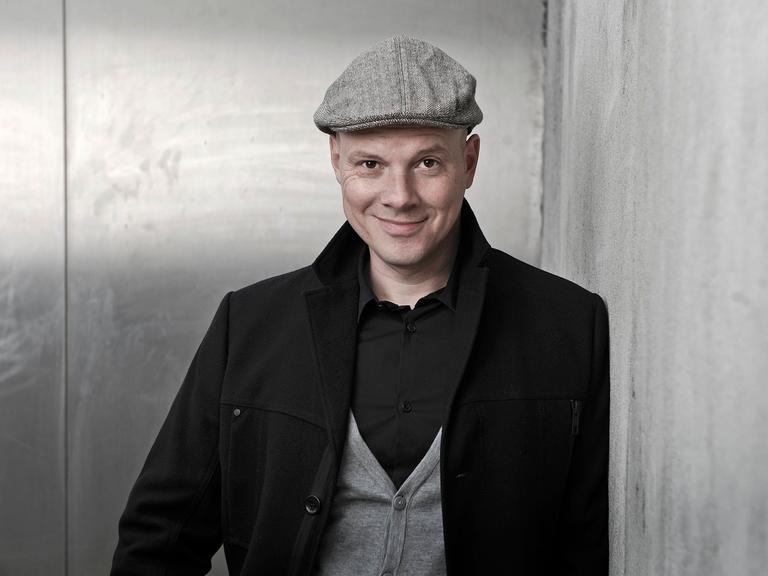 Oliver Hilmes. Er trägt eine schwarze Jacke, graue Schiebermütze und schaut lächelnd in die Kamera
