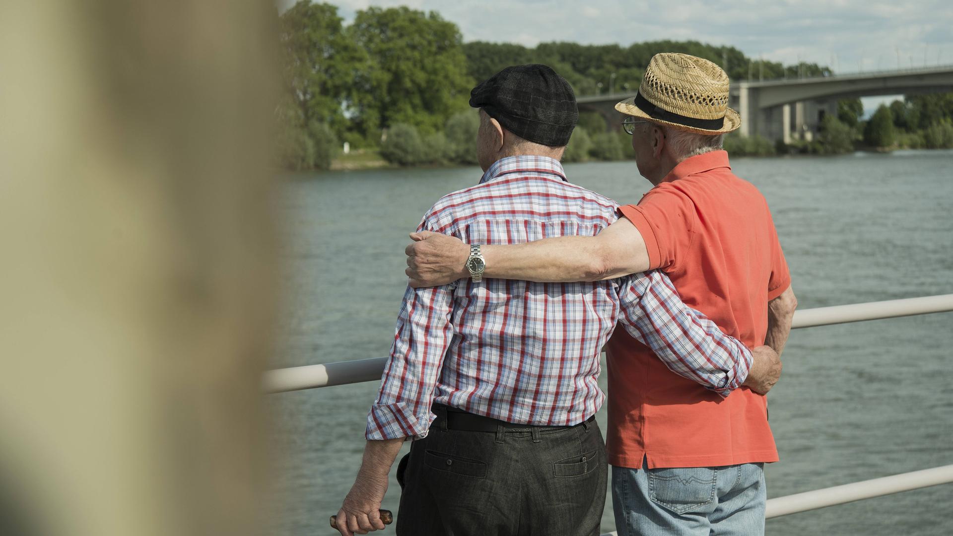 Zwei ältere Männer stehen umarmt an einem Fluss und schauen aufs Wasser.