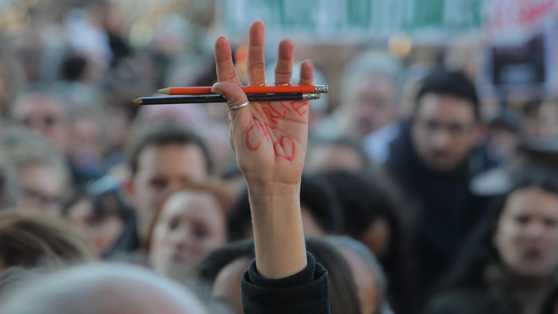 Eine Person in einer Menschenmenge hält ihre Hand nach oben, in der sie zwei Stifte hält. Auf ihrer Hand steht "Charlie" geschrieben und daneben ist ein Herz gezeichnet. 