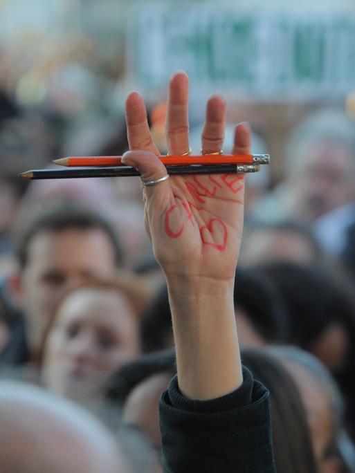 Eine Person in einer Menschenmenge hält ihre Hand nach oben, in der sie zwei Stifte hält. Auf ihrer Hand steht "Charlie" geschrieben und daneben ist ein Herz gezeichnet. 